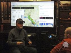 In this Tuesday, March 5, 2019 photo, Iditarod spokesman Chas St. George, left, sits in front of a large screen set up inside a hotel in Anchorage, Alaska, for public updates in the 1,000-mile Iditarod Trail Sled Dog Race. Technology has increasingly made the 47-year-old race more immediate to fans and safer for competitors, said St. George, acting CEO of the Iditarod Trail Committee, the race’s governing board. (AP Photo/Rachel D'Oro)