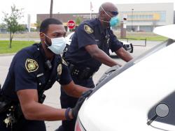 In this April 9, 2020, photo, amid concerns of the spread of COVID-19, Dallas ISD police officers Mylon Taylor, left, and Gary Pierre push a car that ran out of gas while waiting in line for the weekly school meal pick up for students in Dallas. The coronavirus pandemic that has crippled big-box retailers and mom and pop shops worldwide may be making a dent in illicit business, too. (AP Photo/LM Otero)