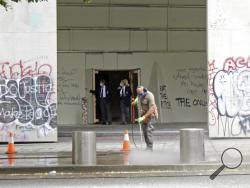 A worker washes graffiti off the sidewalk in front of the Mark O. Hatfield Federal Courthouse in downtown Portland, Ore., on Wednesday, July 8, 2020, as two agents with the U.S. Marshals Service emerge from the boarded-up main entrance to examine the damage. Protesters who have clashed with authorities in Portland, Ore., are facing off not just against city police but a contingent of federal agents who reflect a new priority for the Department of Homeland Security: preventing what President Donald Trump cal