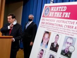 A poster showing six wanted Russian military intelligence officers is displayed as Assistant Attorney General for the National Security Division John Demers, left, takes the podium to speak at a news conference at the Department of Justice, Monday, Oct. 19, 2020, in Washington. Also pictured is US Attorney for the Western District of Pennsylvania Scott Brady, center. (AP Photo/Andrew Harnik, pool)