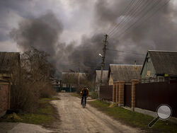 A Ukrainian man rides his bicycle near a factory and a store burning after it had been bombarded in Irpin, on the outskirts of Kyiv, Ukraine, Sunday, March 6, 2022. (AP Photo/Emilio Morenatti)