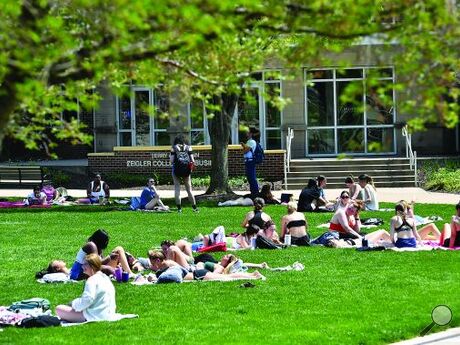 Students in shorts and tank tops lie on blankets on green grass in front of a college building.