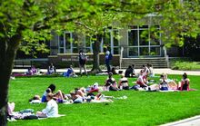 Students in shorts and tank tops lie on blankets on green grass in front of a college building.