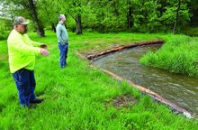 Two men on a green field look at logs protecting the banks of a stream.