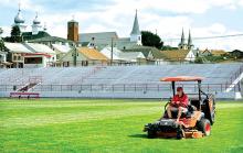 With the church spires of Mount Carmel in the background, groundskeeper Jake Wojcik cuts the grass on the football field in Mount Carmel High School’s Silver Bowl on Friday morning.